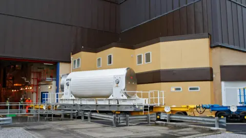 Sellafield A flask standing on a platform outside an industrial building. A group of men in high-visibility clothing can be seen working in the background