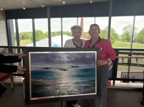 Holly Phillips/ BBC Coby Van Riel and Nicky van der Drift standing indoors by large windows with a scenic green park and tent visible outside. They are smiling and holding a framed painting. The artwork depicts three World War Two bombers flying low over a reflective sea under a cloudy sky at dusk or dawn.