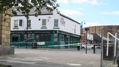 Police tape has been sprung across Shields Road in Byker. The Raby pub is in the background. A police officer stands watch over the cordon.