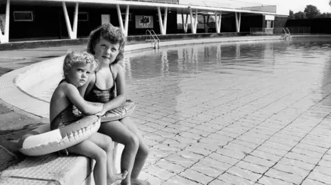 Getty Images Two young girls sit beside the water at Stechford Baths in 1987. Both girls have a rubber ring around them and the smaller girl is resting her head against the older girl.