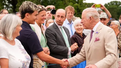 Reuters Young man with dark hair and blond streaks shakes King Charles' hand as other members of the public look on. 