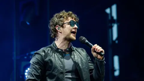 A man with brown curly hair, sunglasses and a black leather jacket holding a microphone against a dark background