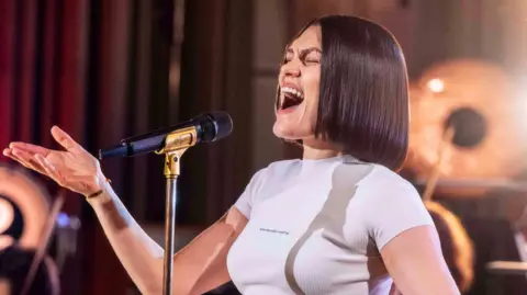 Jessie J sings into a mic with an orchestra behind her. She has a black bob and wears a white T-shirt 