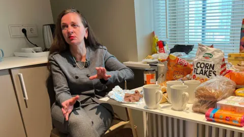 A woman in a grey suit sits next to a kitchen table loaded with food
