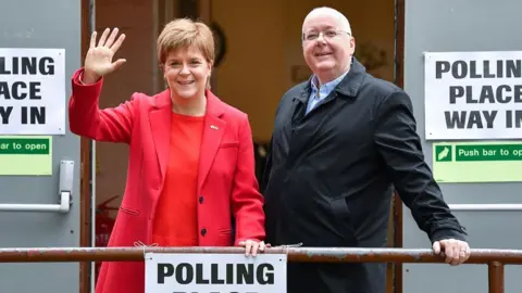 Getty Images Nicola Sturgeon, who has short fair hair, and Peter Murrell, who is bald, standing smiling outside a polling station while leaning on a hand rail. Sturgeon, on the left, is wearing a pink jacket and red top. Murrell is wearing a dark overcoat. 
