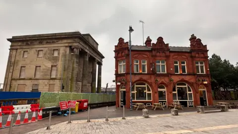 Supplied The image shows two buildings, one a weathered stone structure with boarded-up windows and the other a red brick pub with outdoor seating. Construction barriers and traffic cones are visible in the foreground. 
