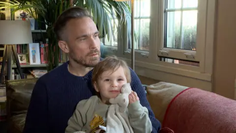 The Dad holds his son who clutches a teddy to his face. They sit on a sofa with a plant in the background.