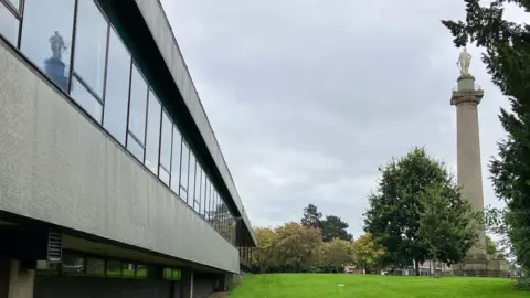 A side image of Shropshire Council's headquarters, Shirehall, on the left, and Lord Hill's statue on top of a tall stone column on the right. There is greenery and trees in the background.