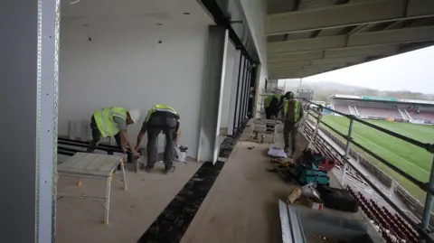 Northampton Town Football Club/ Peter Norton Two men in green high visibility jackets and white hard hats measuring a metal beam standing on the balcony of what will be a box area of the new East Stand at Northampton Town Football Club. 