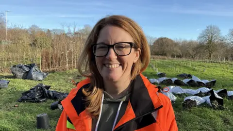 Alex Morel stands in a field, with lots of newly planted trees in the background. There are also bagged trees on the floor waiting to be planted. Alex has long brown hair, black framed glasses and a bright orange rain coat. 