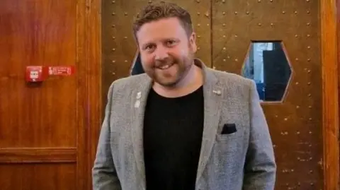 Alex Winning smiles at the camera while standing near a double door in a wood-panelled room. He has 