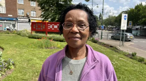 Norma Thompson in Wednesfield town centre. There are some shops and a car behind her. She has black hair and is wearing a jumper and purple fleece. She has a necklace on and is wearing glasses.