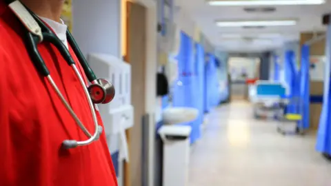 A stock image of a hospital ward. The image shows an empty ward with several bays with blue curtains, which are blurred in the background. In the forefront of the image is a man in bright red scrubs with a stethoscope around his neck. 