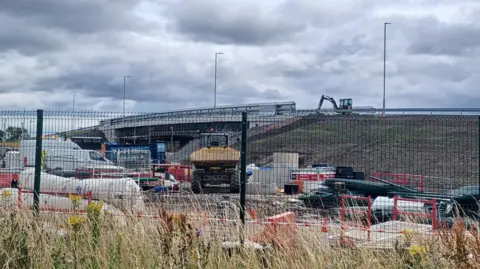 Northumberland County Council Taken from behind a wire fence, you can see lots of construction work on a road bridge above a rialway line. A digger is at work on the structure itself 