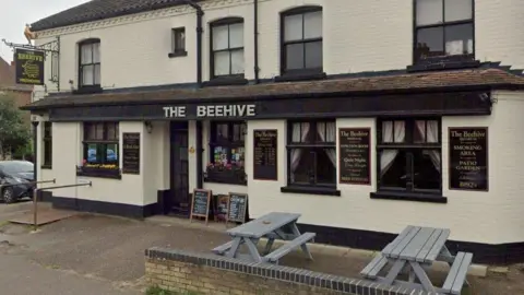 Google Exterior of The Beehive pub. The walls are painted cream and the window and door frames are painted black. Pub signage is on the walls and there are grey benches out the front.