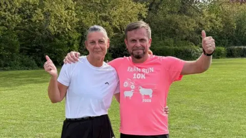 Chris Grubb A couple in their fifties stand next to each other with an arm around each other. Clair has her grey hair in a ponytail and wears a white t-shirt. Chris has a neon pink running top on, and had brown hair and a brown goatee. Both give a thumbs up to the camera.
