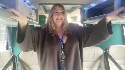 Paul Burnell/BBC Janey Parish is standing in the aisle of a coach. She has shoulder lenght blonde hair and is wearing a blue lanyard.