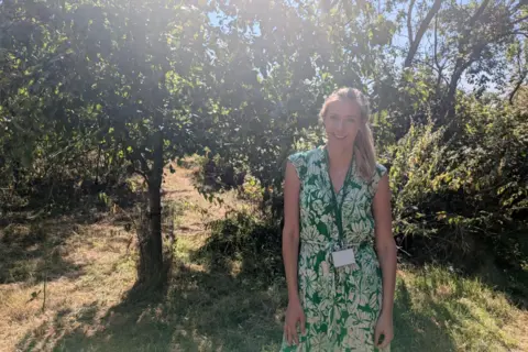 A woman with blonde hair in a ponytail is wearing a green and white floral print dress and smiling as she stands on dry grass in an orchard with green trees behind her and dappled sunlight breaking through the foliage.