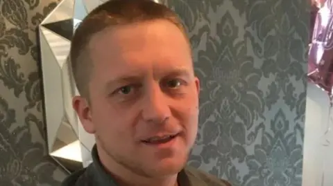 South Yorkshire Police Bradley Hollis, smiling to camera. He has short fair hair. Behind him is patterned wallpaper, a mirror decoration and a party balloon. 