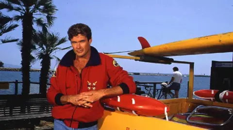 David Hasselhoff in a red coat leaning on a yellow truck.