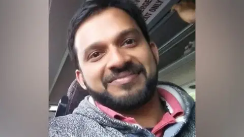 Spindrift A man with a beard and a goatee smiling into the camera. He has short dark hair, and appears to be seated on a train.