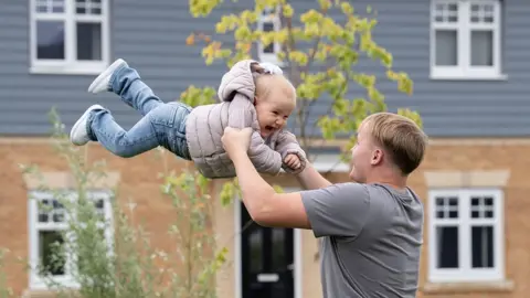 NEYDL Alex Tarling, who's wearing a grey T-shirt and has short, blond hair, is swinging his small daughter around in the air. She's wearing jeans and a puffy coat and is smiling.