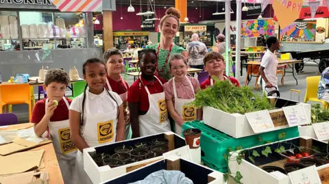 Alex Moss/BBC A group of six schoolchildren -three boys and three girls - standing with their female teacher behind a stall displaying boxes of fruit and vegetables. 