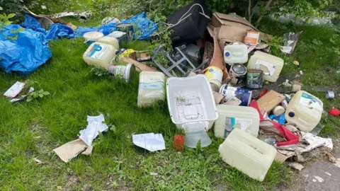 A pile of rubbish dumped on the roadside in Kirklees. The pile includes large plastic containers and bags. 