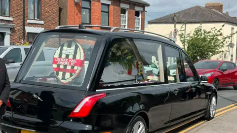 A white coffin covered with white flowers and a wreath of the Lostock Hall Junior Football Club logo are in the rear window of the black hearse at Kerry Coombes funeral.