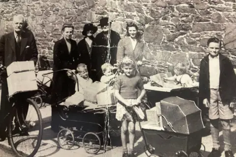 David Isherwood A black and white World War Two era photo of a family carrying packages wrapped with paper and string - standing in front of a granite wall. There are two older adult men - one on a bicycle with packages on the front. Next to them three adult women and three toddlers, a young girl holding a pram full of packages, and a young boy in shorts,