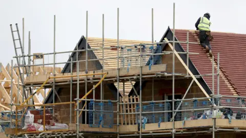 A construction of brick houses. There is scaffolding on the side of the property roofs and a person in a hi-vis jacket working on the roof of one of the buildings closest to the camera on the right of the frame.