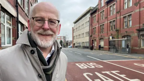 Mark Jewell: a balding, white man with a white beard. He is smiling, wearing a grey trench coat, green scarf and brown-rimmed glasses. He is standing on Corporation Street in front of the bus gate marked on the road in big white capital letters. There are buildings on either side of the road.
