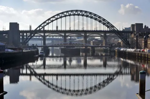 Jeff Overs/BBC A wide view of the Tyne Bridge from the river Tyne. It is a metal bridge, painted green. Several other bridges can be seen behind it. The silhouettes of the bridges are reflected in the water below.