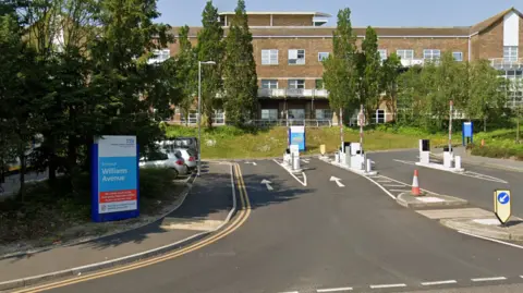 Google A screenshot from Google street view of the road entrance to a hospital. There's a barrier system on the road, a blue and white sign on the left hand side and a brick building in the background. It's a sunny day and the sky is blue