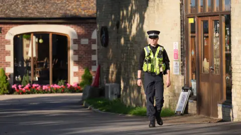 PA Media A single uniformed police officer walking past a brick building. They were a black hat, yellow hi-vis jacket with a black top, trousers and shoes. 