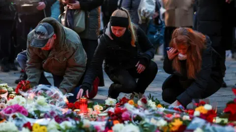 Reuters Three people - a man and two women - lay tributes near the crime scene. In the foreground you can see flowers of different types and colors as well as candles in glass containers.