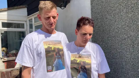 A man and a woman stand outside a house wearing matching T‑shirts printed with a photo and the hashtag “#justiceforcarris.”
