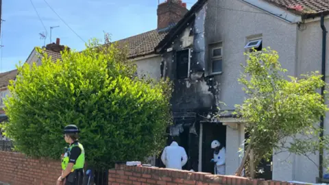 A fire-damaged house on a residential street. A police officer is standing in front of the property and two forensic officers are standing in the entrance. The entrance to the house and an upstairs window are burnt out. 