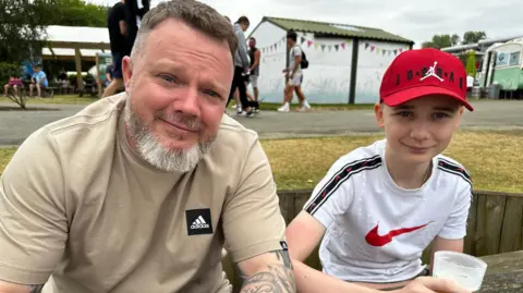 A man with short brown hair and a grey beard, wearing a beige Adidas t-shirt, sits at a picnic table next to a young boy wearing a white Nike t-shirt and a red baseball cap. The boy is holding a drink and both are smiling at the camera.