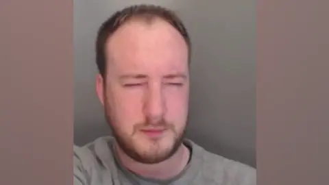 Cleveland Police A custody photo of Nathan Henderson who has short brown hair and a beard and is wearing a grey shirt. His eyes are closed and he is leaning to the left of the frame.