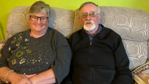 Joyce's children Jennifer and James sit on a grey sofa, against a bright green backdrop. Jennifer wears a grey jumper with sequinned flowers, and James wears a black zip-through jumper and a blue and white shirt.