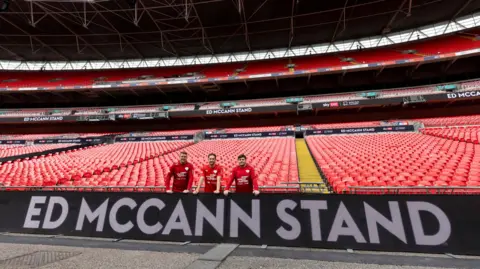 Sky Bet BHF Every Minute Matters Three men in red shirt stand in an empty football stadium. At the front of the stand is a sign that reads: Ed McCann Stand