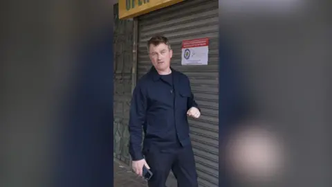 A man wearing a black shirt and jeans stands in front of a shuttered shop