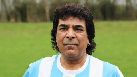 A man resembling Diego Maradona is stood on a football pitch. He is dressed in a blue and white football shirt, has thick black hair and wears an earing.