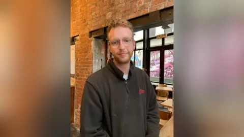 Steven is standing in his café. He wears clear glasses and has fair hair and a short beard. He's wearing a black hoodie. The café has exposed brick walls and brown wooden tables and chairs.