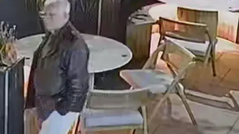 CCTV footage of a man standing beside a tall, dark planter that contains a bushy plant. He is wearing a dark jacket over a top and light‑coloured trousers, and is positioned at a slight angle, facing toward the right side of the image. Behind him is a round white table with multiple chairs placed around it. Additional chairs are visible further back, some tucked under tables and others slightly angled.