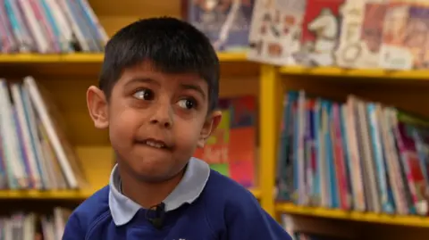A boy with dark hair is looking towards the right on the photo. Books on shelves are blurred behind him.
