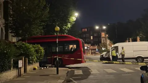 A red bus is crashed into the front of a building. People stand behind it next to a white van.