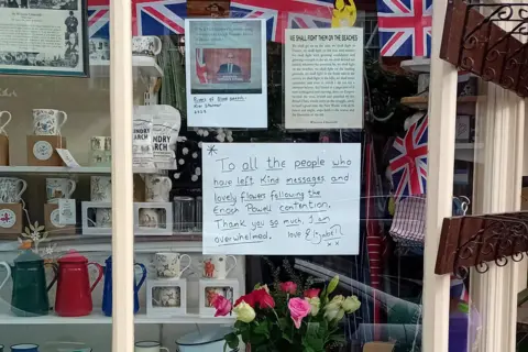 Elizabeth Griffiths A shop window with cups, teapots, flowers and Union flags on display.
There is also a message which reads "To all the people who have left kind messages and lovely flowers following the Enoch Powell contention Thank you so much I am overwhelmed."