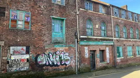 BBC/Victoria Scheer A long, weathered brick building with multiple rows of windows, many of which have peeling turquoise paint on the frames and shutters. Graffiti covers part of the lower wall, including a large, bold piece in white, black and pink. Several windows are boarded up or damaged, and pipes run vertically along the façade.